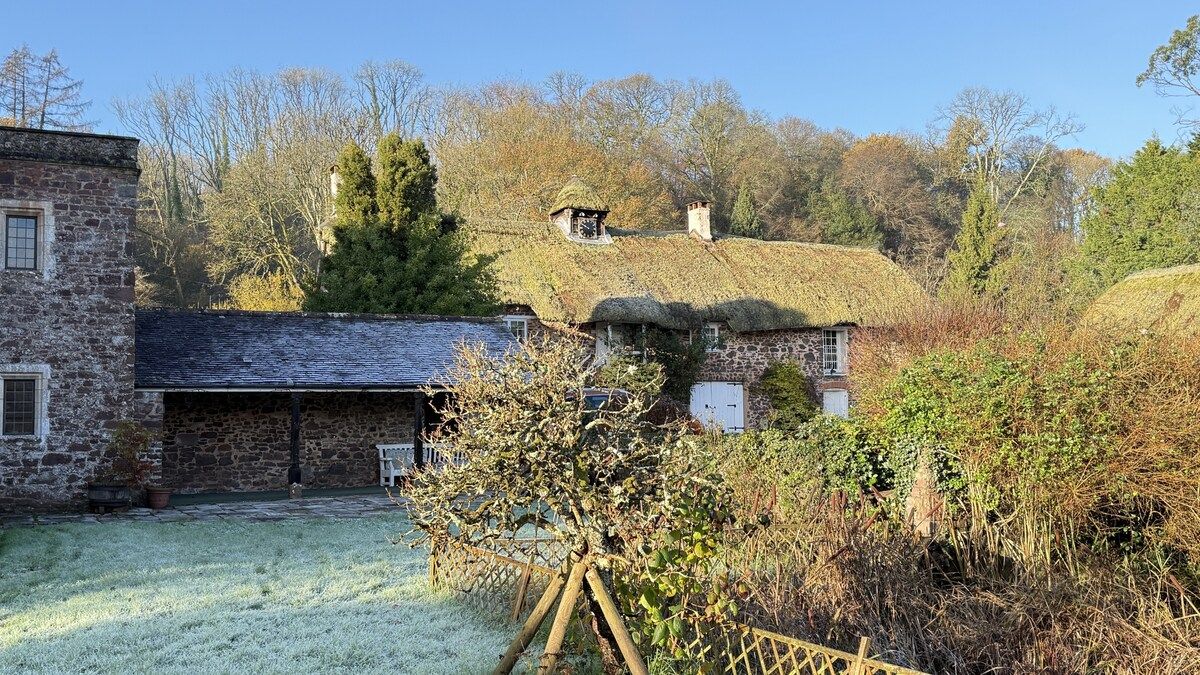 A stone building with a thatched roof, a fenced yard, and a tree in the foreground.