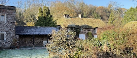 A stone building with a thatched roof, a fenced yard, and a tree in the foreground.