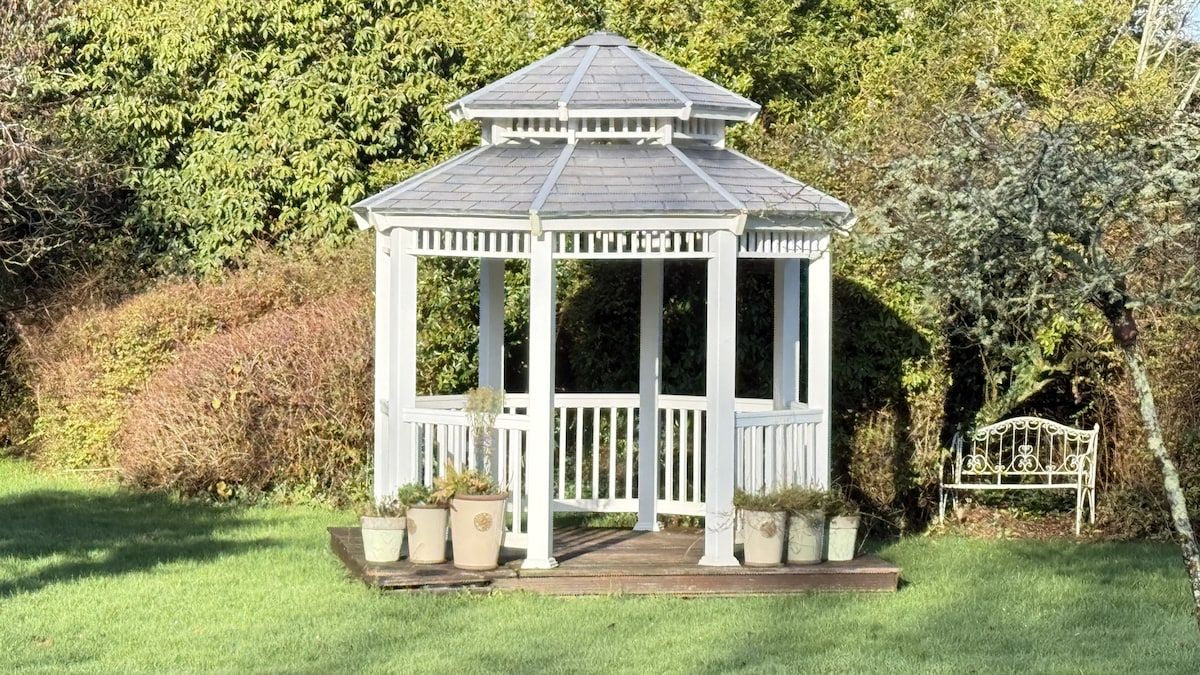 A gazebo with a white frame and a grey roof, surrounded by greenery.