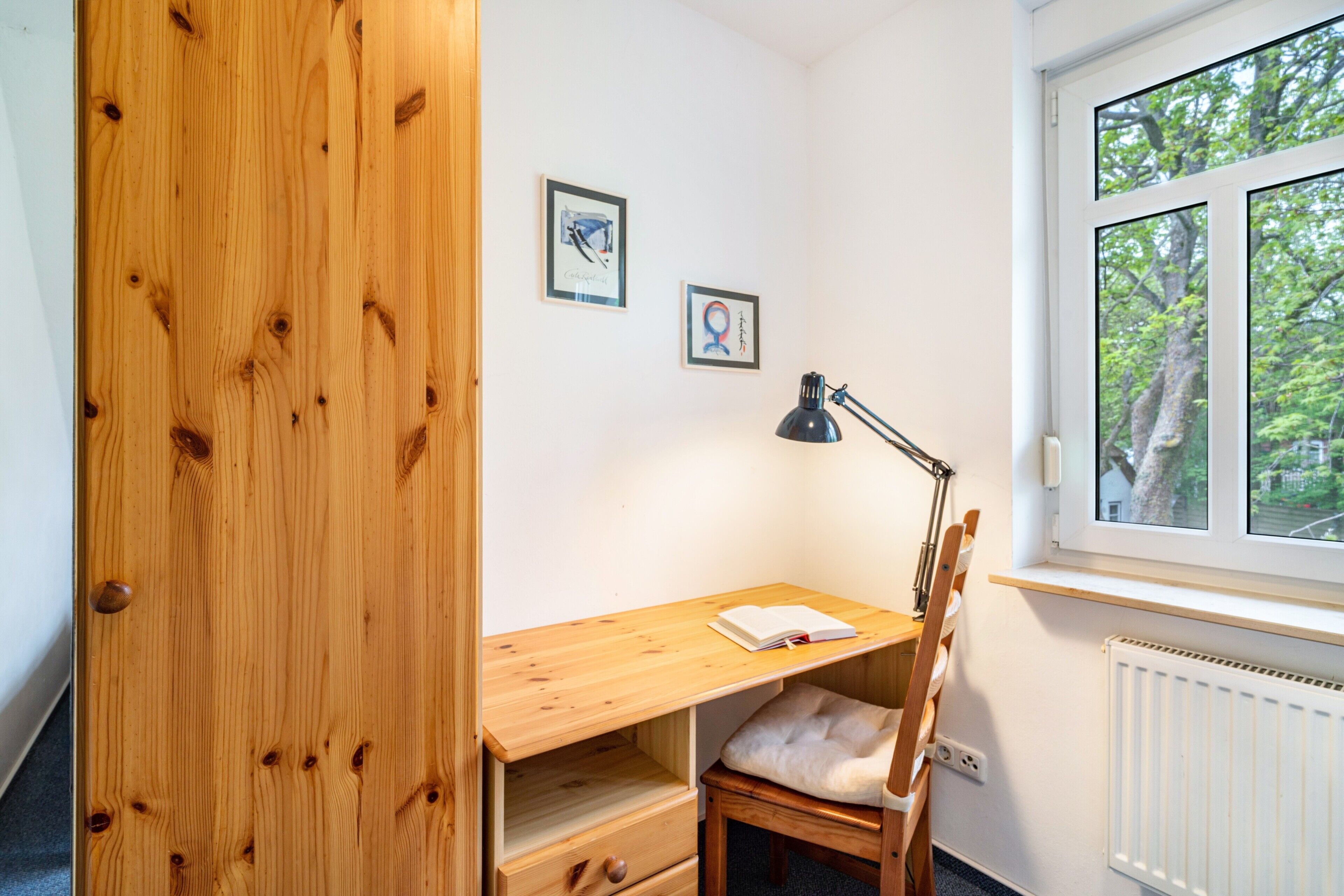 Een kamer met een houten bureau, een stoel, een raam met uitzicht op bomen, en twee ingelijste foto's aan de muur.