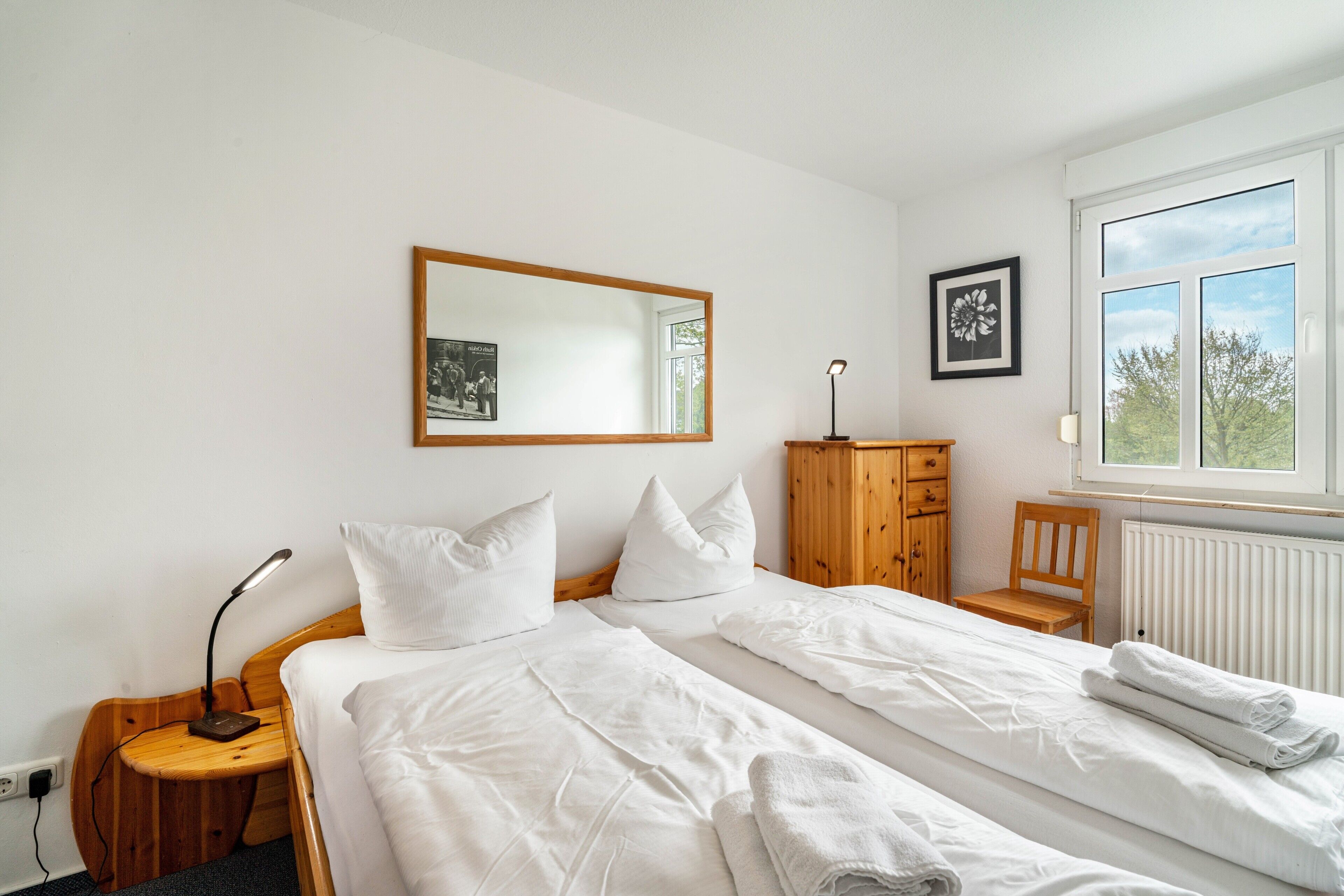 A bedroom with two beds, a wooden cabinet, a desk with a lamp, and a window with a view of trees.