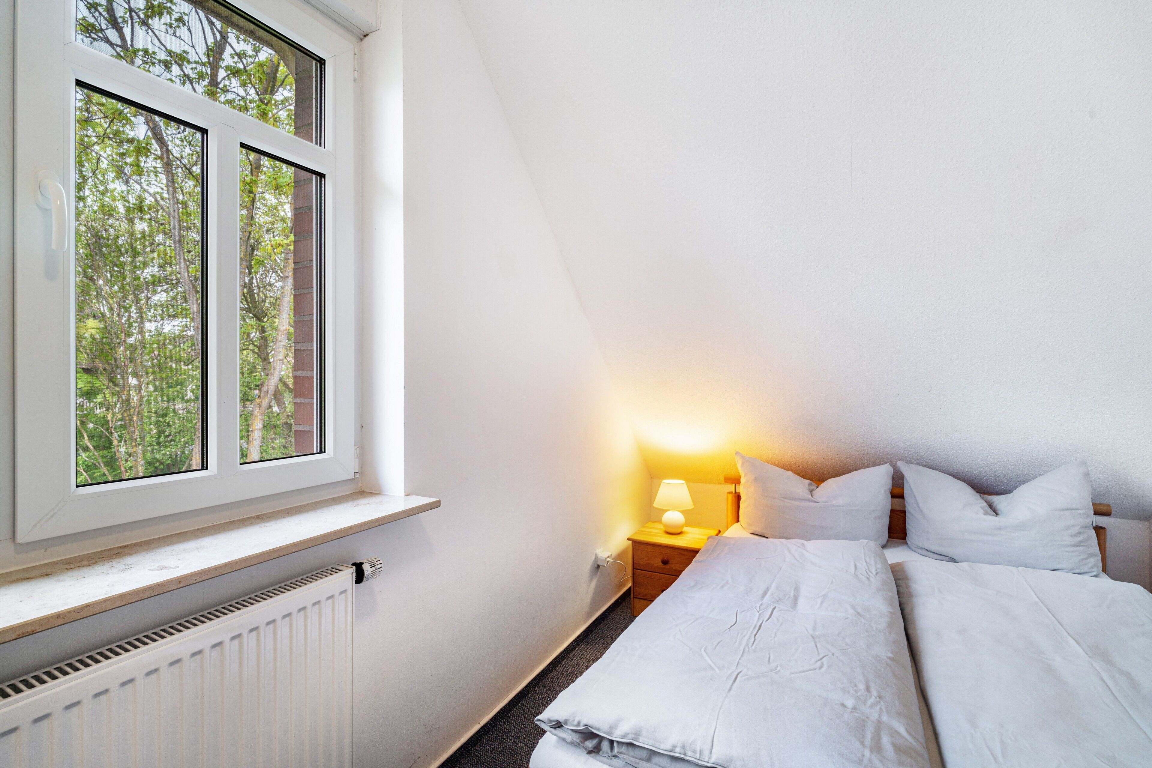 A bedroom with a window, a bedside table with a lamp, and a bed with pillows.