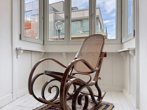 Un fauteuil à bascule se trouve dans une pièce aux murs blancs, d’où l’on peut admirer la vue sur les bâtiments à travers de grandes fenêtres.