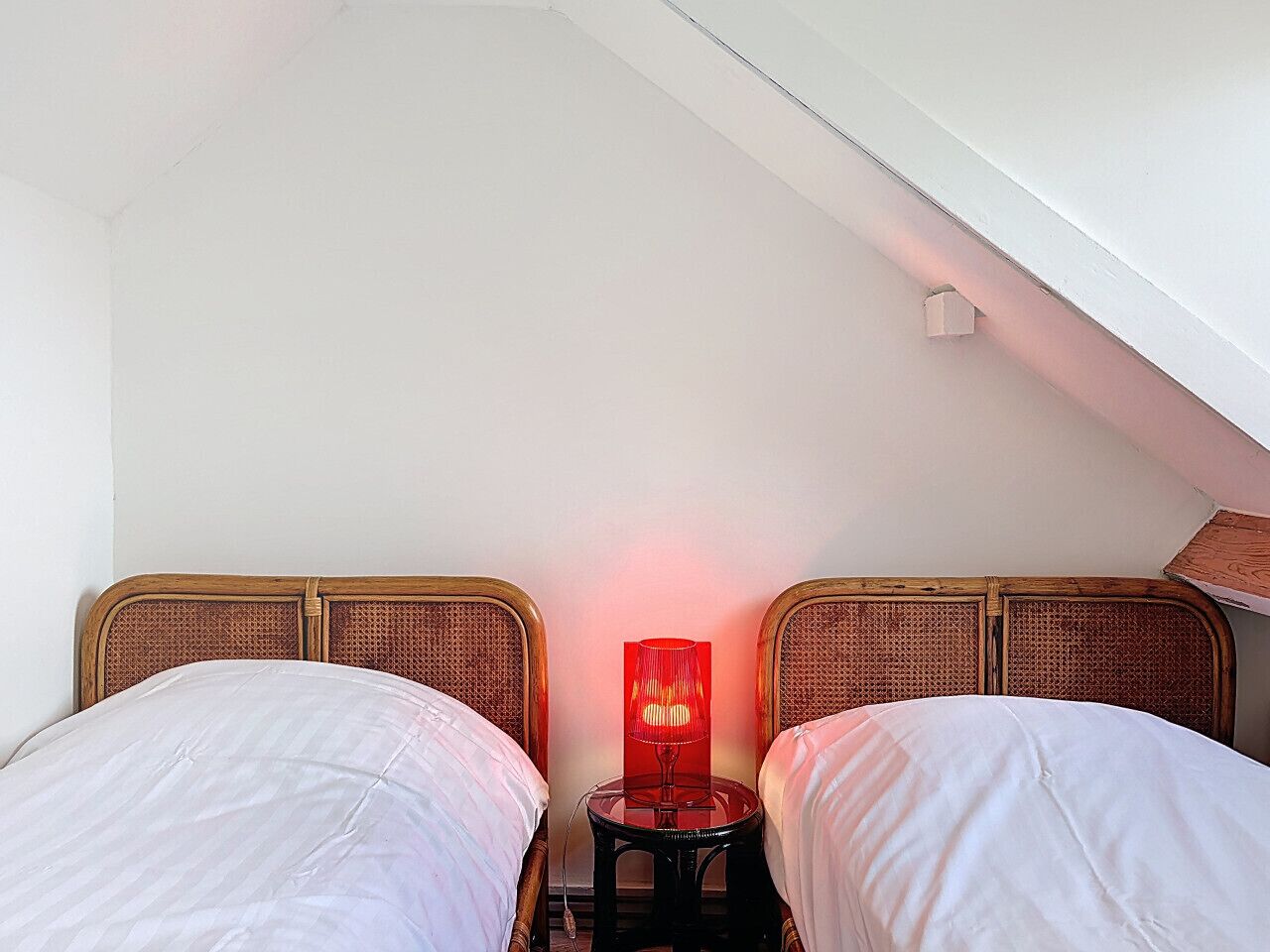 Een slaapkamer met twee eenpersoonsbedden, een nachtkastje met een rode lamp en een schuine balklaag.