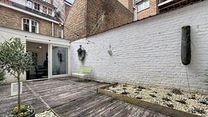 A courtyard with a wooden deck, a bench, a planter with flowers, and a white brick wall. - Maison No Catiau - De Panne (De Panne)