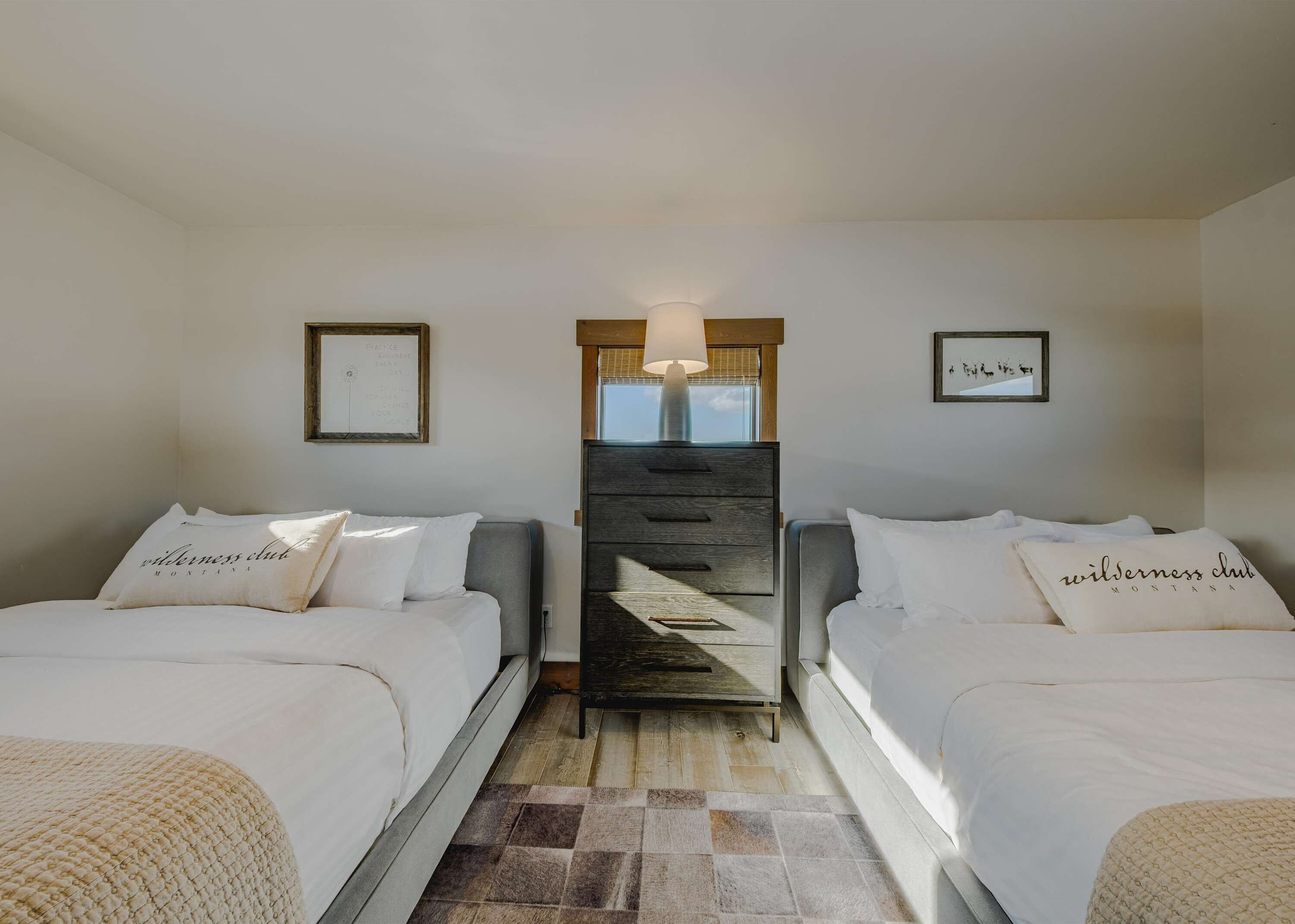 A hotel room with two beds, a wooden dresser, a lamp, and framed pictures on the walls.