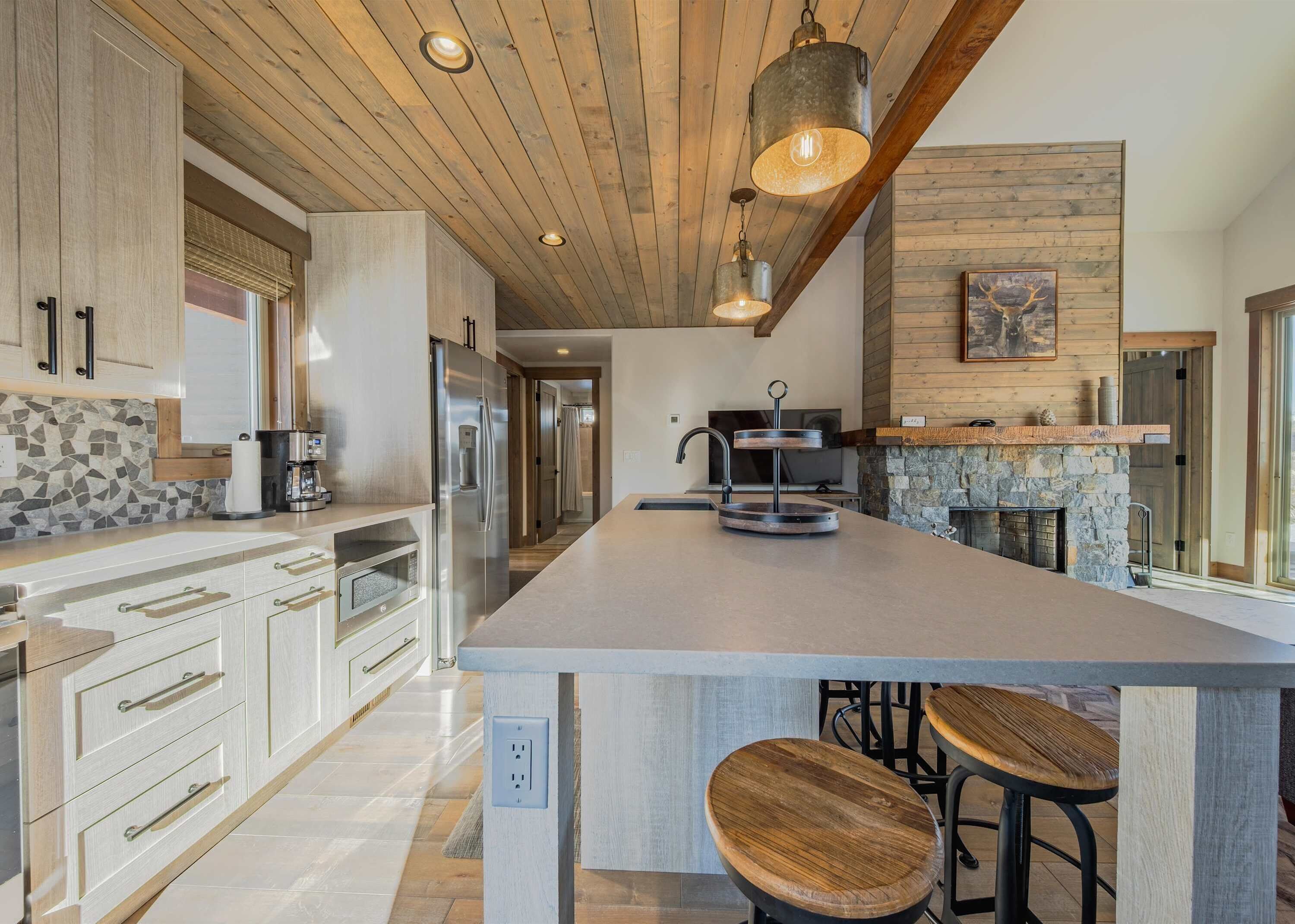 A spacious kitchen with a large island, wooden ceiling, and stone fireplace.