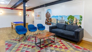 Une pièce avec une table de billard, des chaises bleues, un canapé gris et un tapis coloré.
