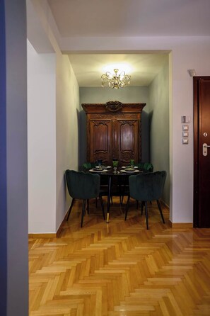 Un espace repas comprenant un buffet en bois, des chaises recouvertes d’un tissu vert et un parquet à motifs en chevrons.