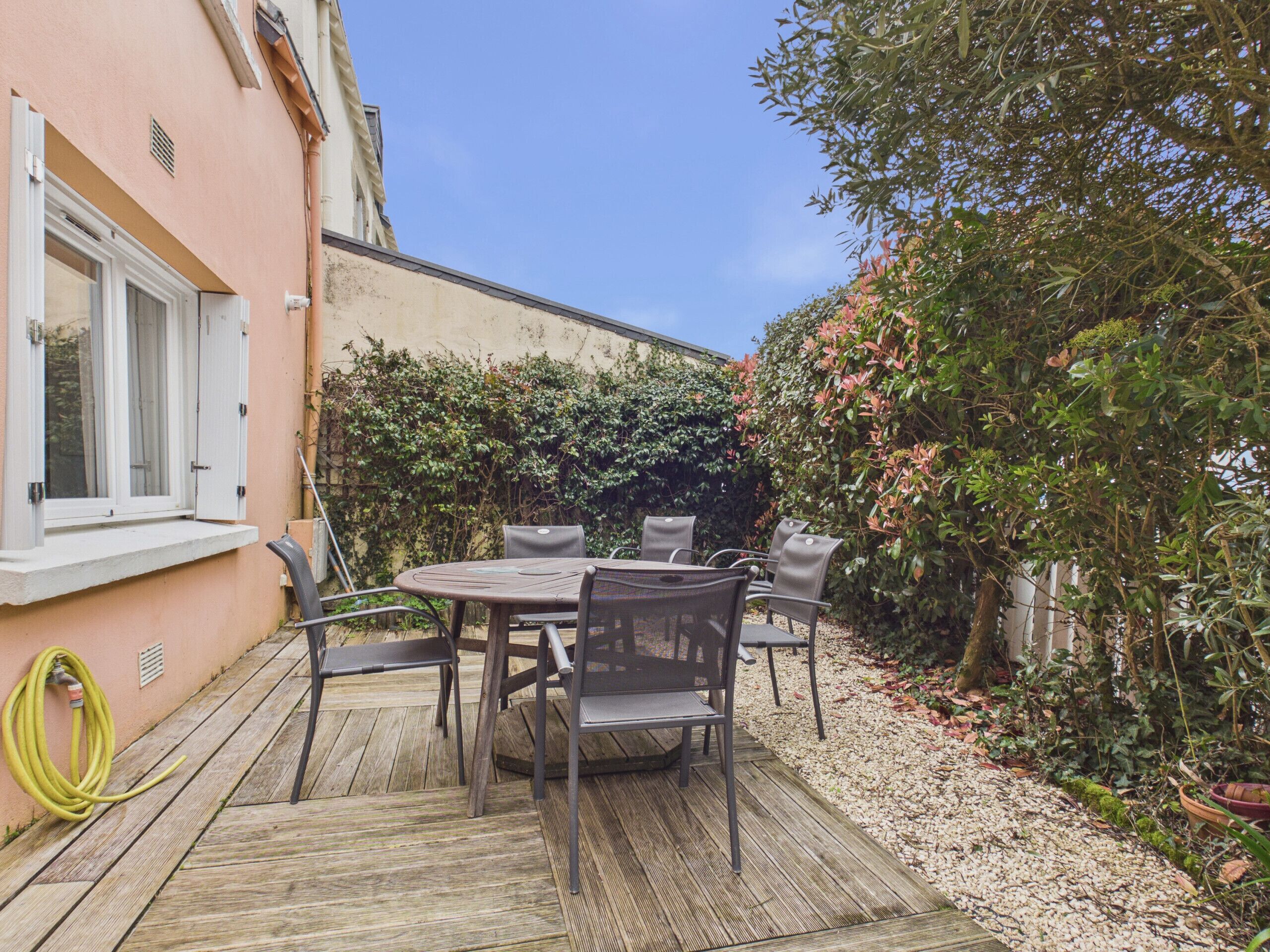 Eine Terrasse im Freien mit Tisch und Stühlen, einem Gartenschlauch und einem Gebäude mit einem Fenster.