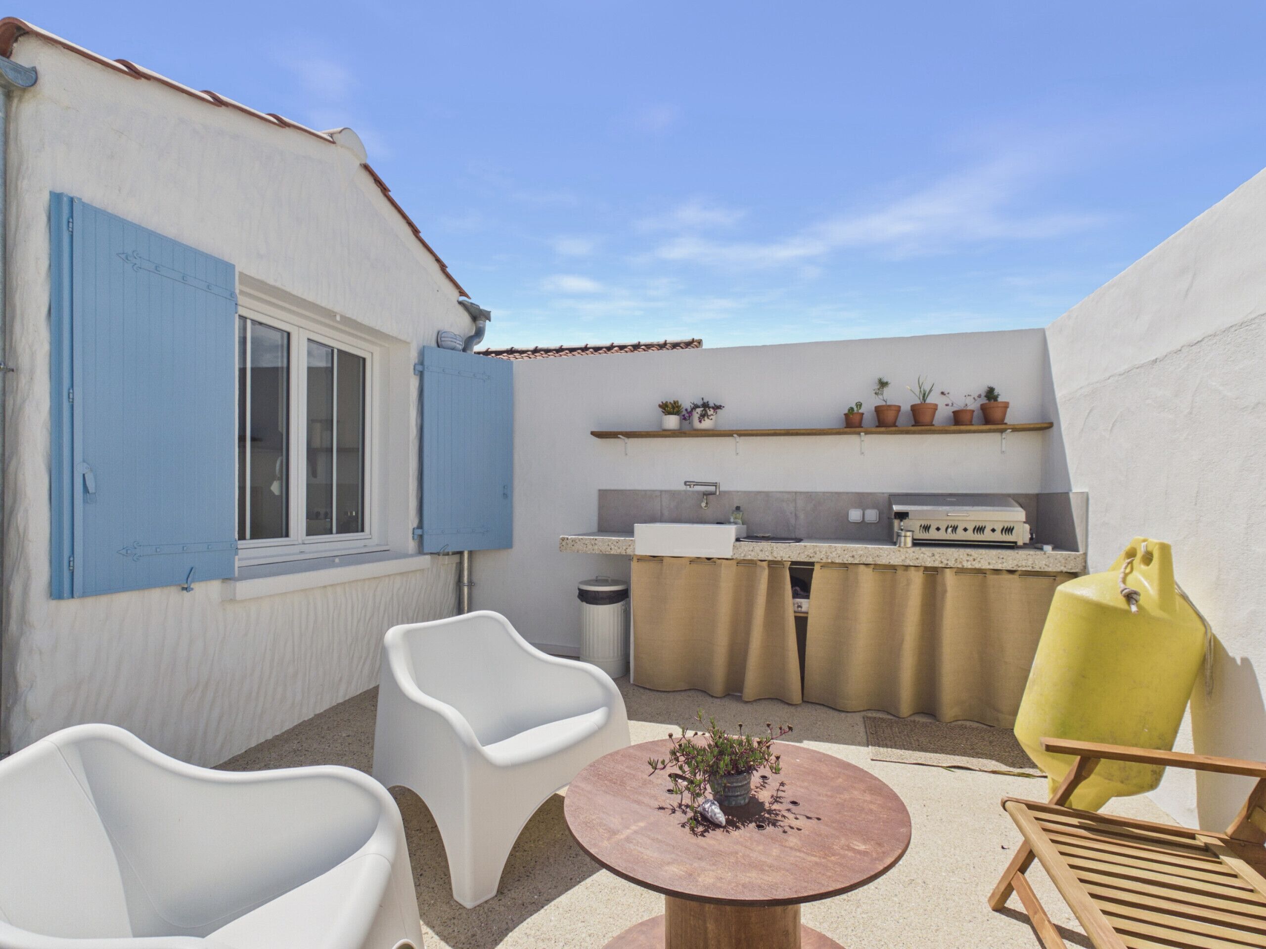 A patio with white chairs, a round table with a plant, a yellow chair, and a kitchen area with a sink and potted plants.