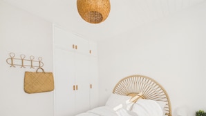 Una camera da letto con un letto, un armadio bianco, un cesto appeso, una testiera in rattan e un comodino con una lampada e una sveglia.