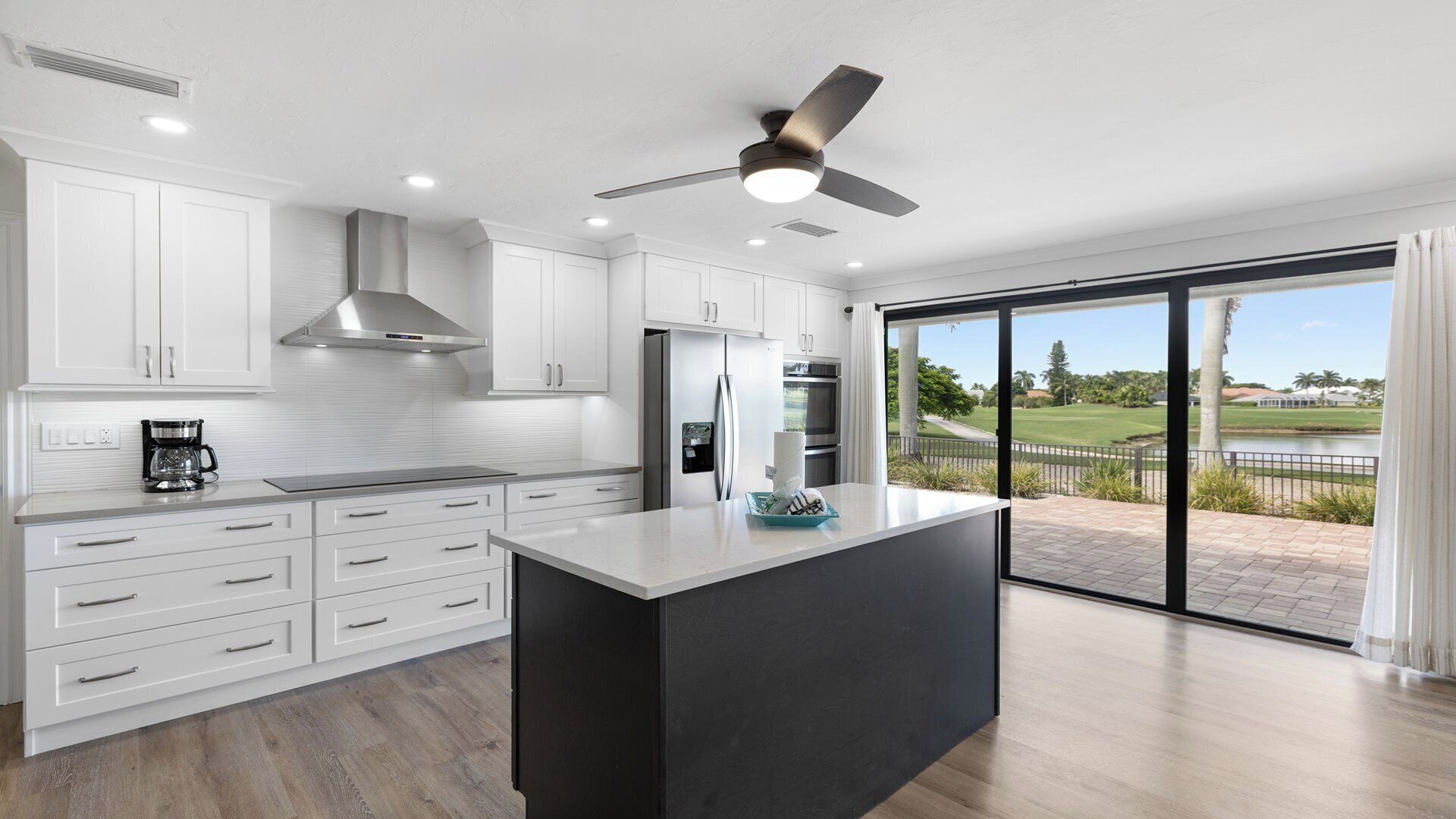 A modern kitchen with white cabinetry, a black island, stainless steel appliances, and a view of a golf course through large sliding glass doors.