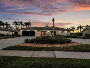 A modern house with a garage, a well-maintained lawn, and palm trees in the background. - Stunning five-bedroom, 3 full bathroom home with views of lake and golf course (Marco Island)