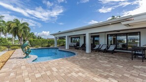 A swimming pool with a unique sculpture, a covered patio area with lounge chairs, and a view of the surrounding landscape. - Stunning five-bedroom, 3 full bathroom home with views of lake and golf course (Marco Island)