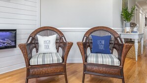 Two wicker chairs with striped cushions and pillows labeled 'HIGH TIDE' and 'LOW TIDE' in a room with wooden flooring and a television on the wall. - Brass Rail 310 | Pool View Balcony, Elevator, Close to Beach (Tybee Island)