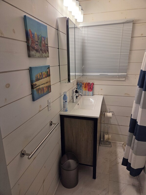 Een badkamer met een witte wastafel, spiegel en handdoekrek. Twee schilderijen hangen aan de muur, er is een prullenbak en een raam met jaloezieën.