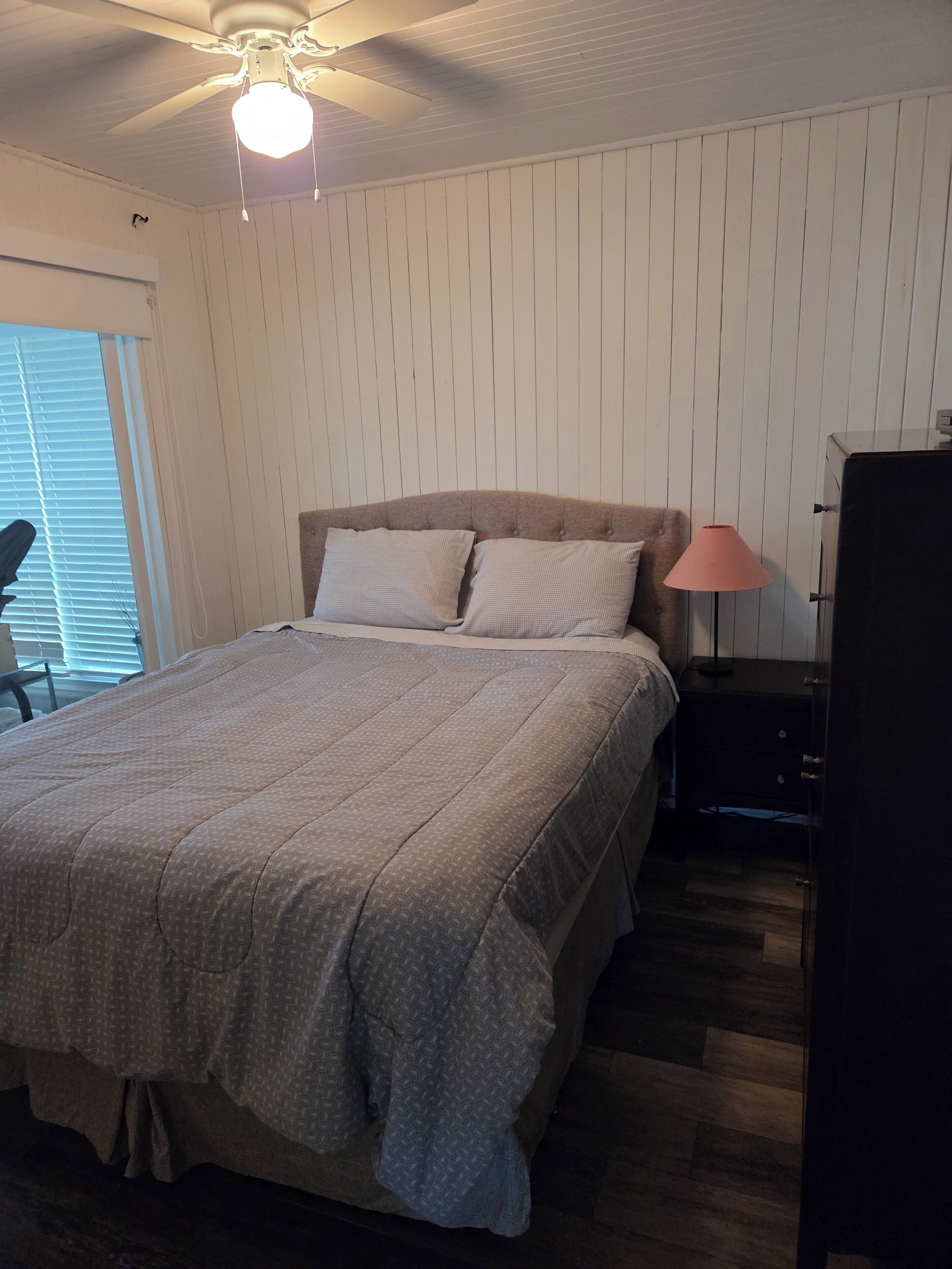 Un dormitorio con cama, ventilador de techo, mesita de noche con lámpara y cómoda.