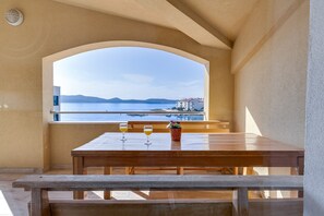 Una mesa de madera con dos copas de vino y una planta en maceta, frente a una vista del mar y edificios lejanos.