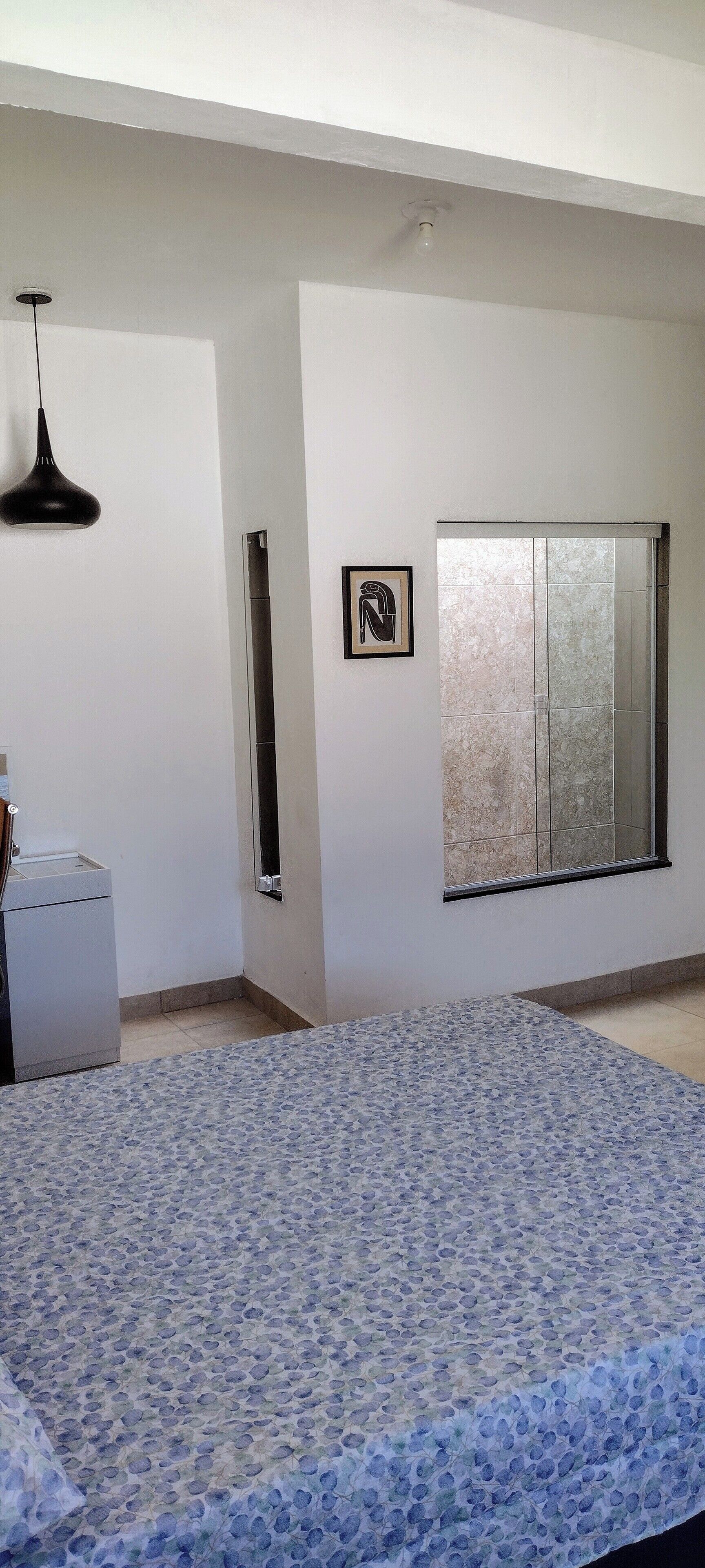 A room with a patterned carpet, a window with a view, a wall-mounted picture, and a ceiling light fixture.