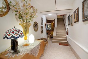 A hallway with a staircase, a table with a lamp, a vase with flowers, and framed artwork on the walls. - Tiepolo – Elegant Room in Central Venice near Biennale and St. Mark’s Square (Venezia)
