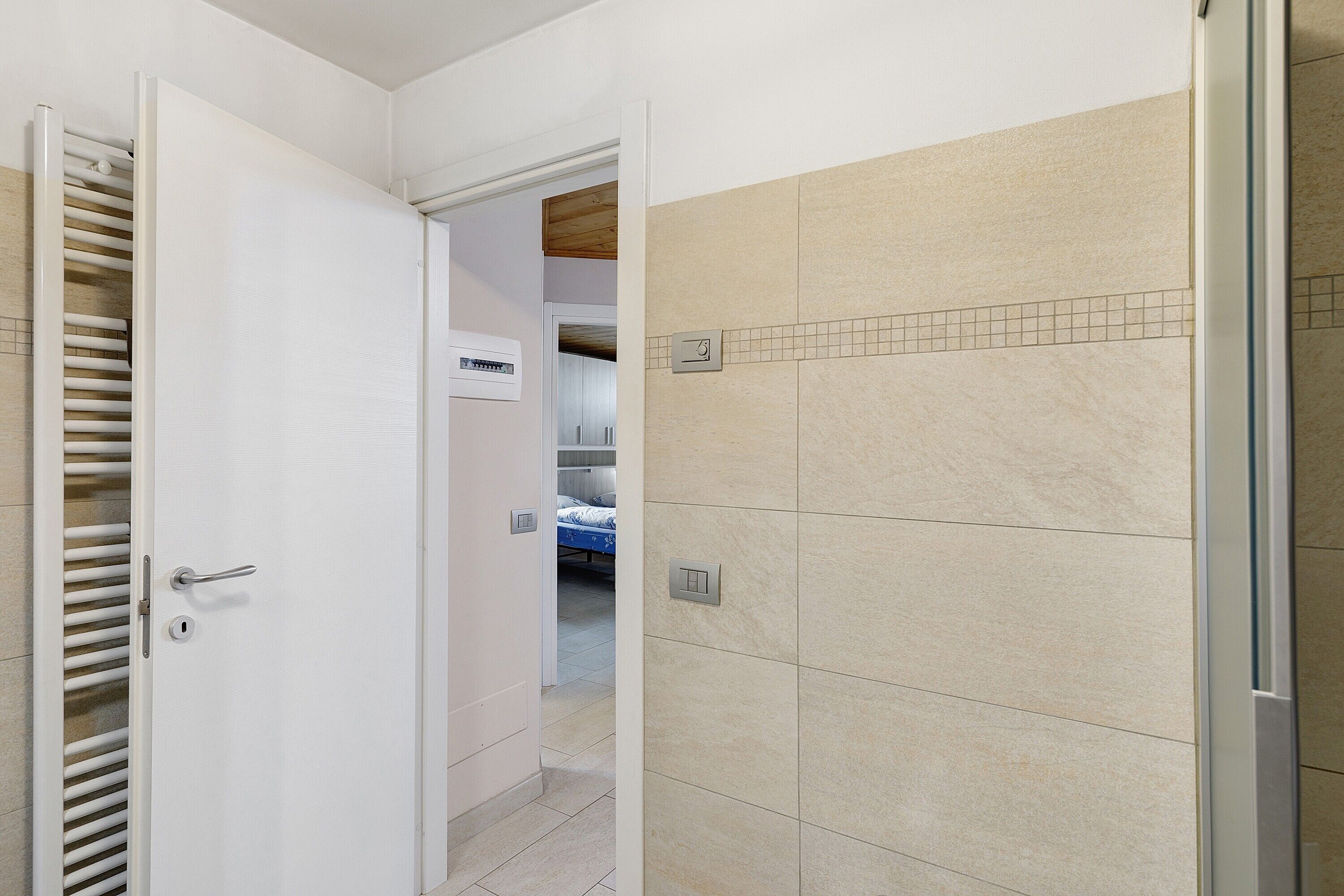 Un couloir avec une porte blanche, des murs recouverts de carreaux beiges et un lit visible au fond.