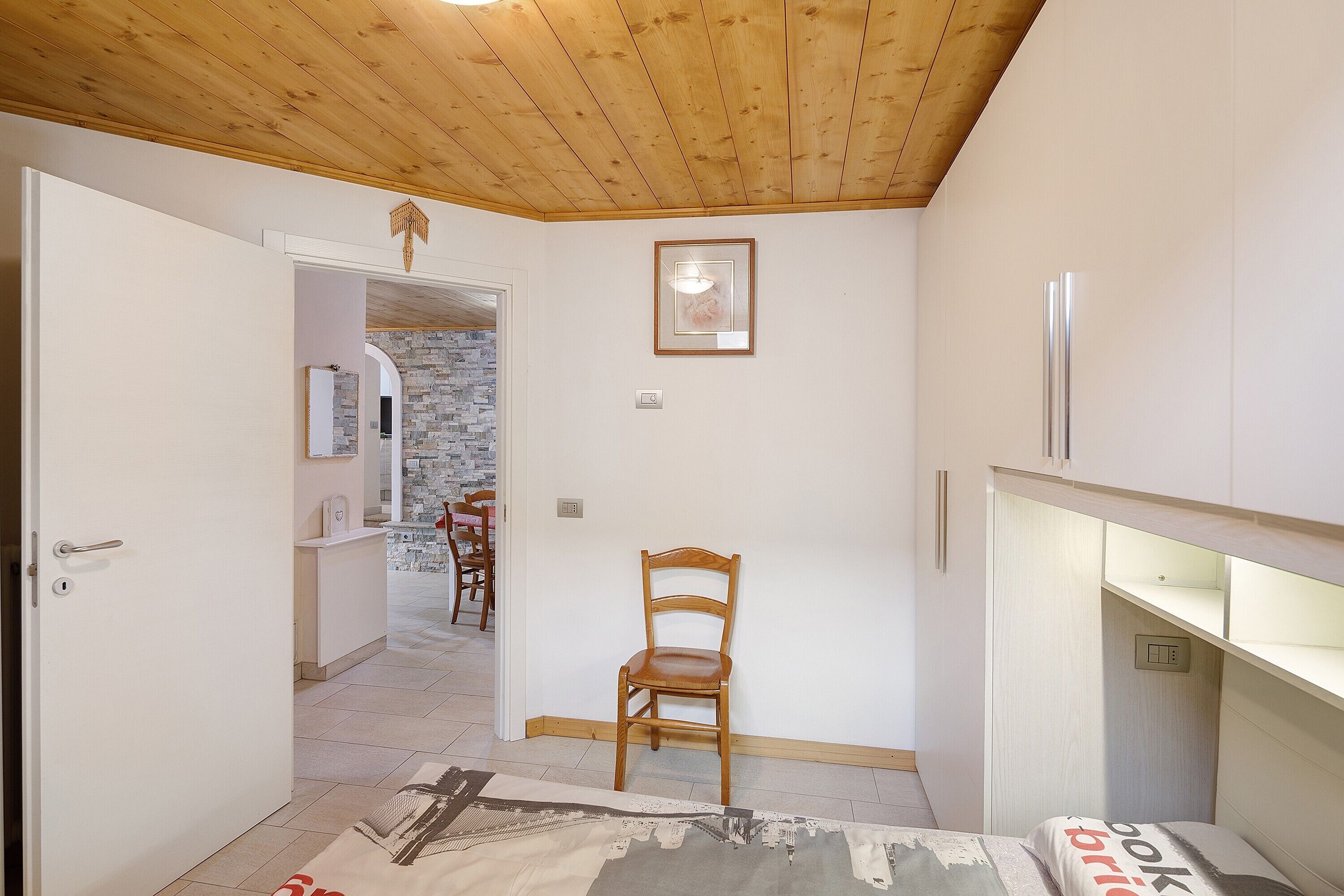 A room with a wooden ceiling, a wooden chair, a bed with a pillow, a white door, and a stone wall in the background.