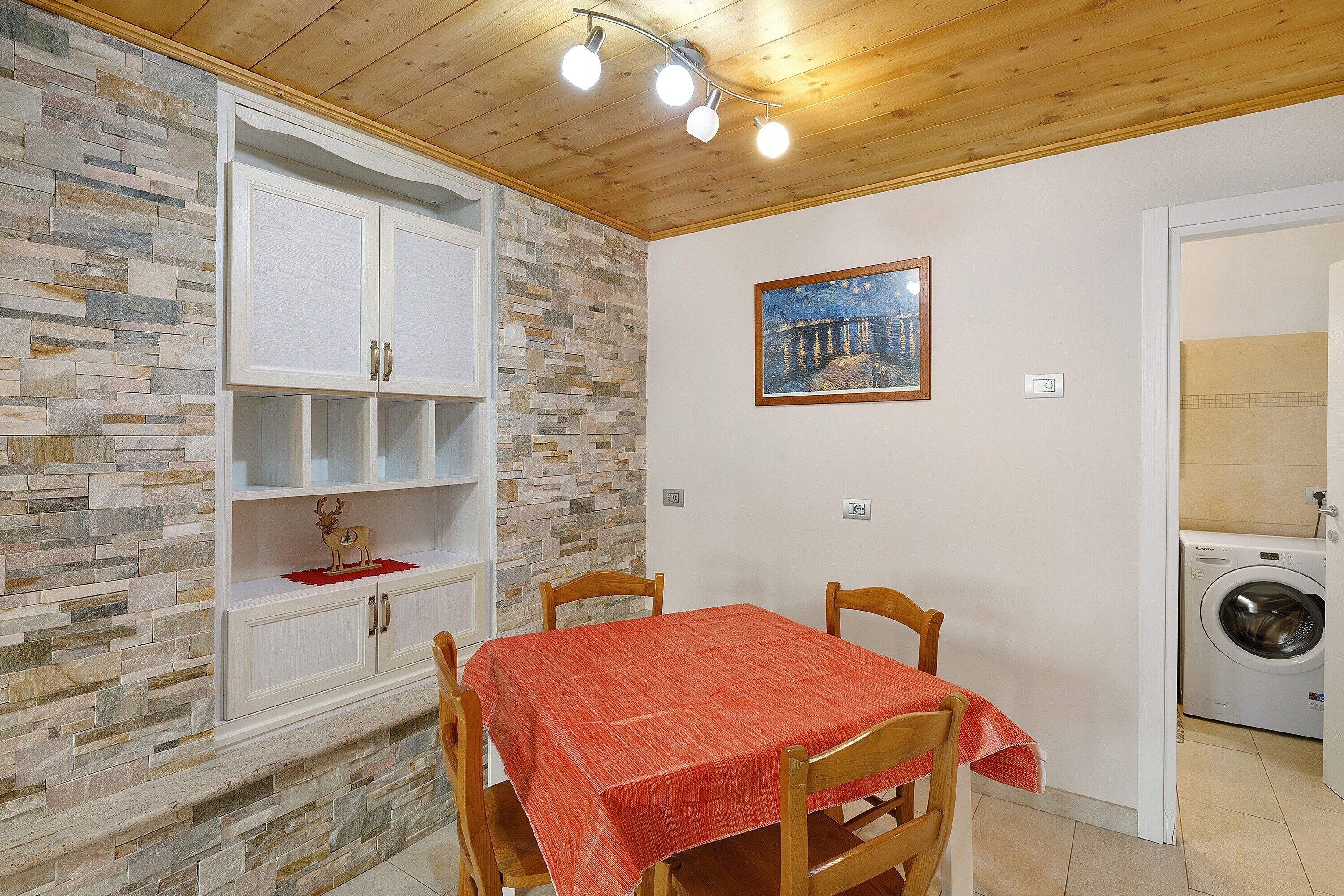 Una habitación con pared de piedra, techo de madera, una mesa con mantel rojo, sillas de madera, un armario blanco y una lavadora.