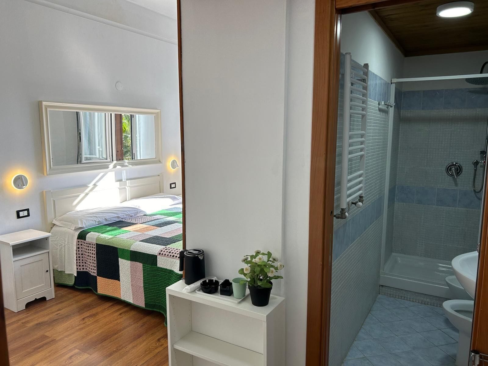 A bedroom with a bed, bedside table, nightstand, and a bedside lamp. There is a bathroom with a shower, sink, and toilet.
