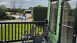 A covered outdoor patio with a round table and chairs, a green building with a sliding glass door, and a view of a parking lot and greenery. - 8 birth Diamond rated Caravan, en-suite, with decking, Parkdean, Skegness (Skegness)