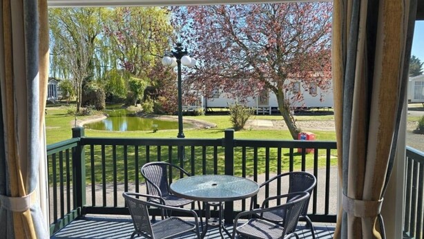 Een overdekt buitengebied met een tafel en stoelen, uitzicht op een vijver en bomen, en een houten terras.