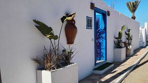 Un bâtiment blanc avec une porte bleue, des plantes en pot et un chemin bordé de carreaux.