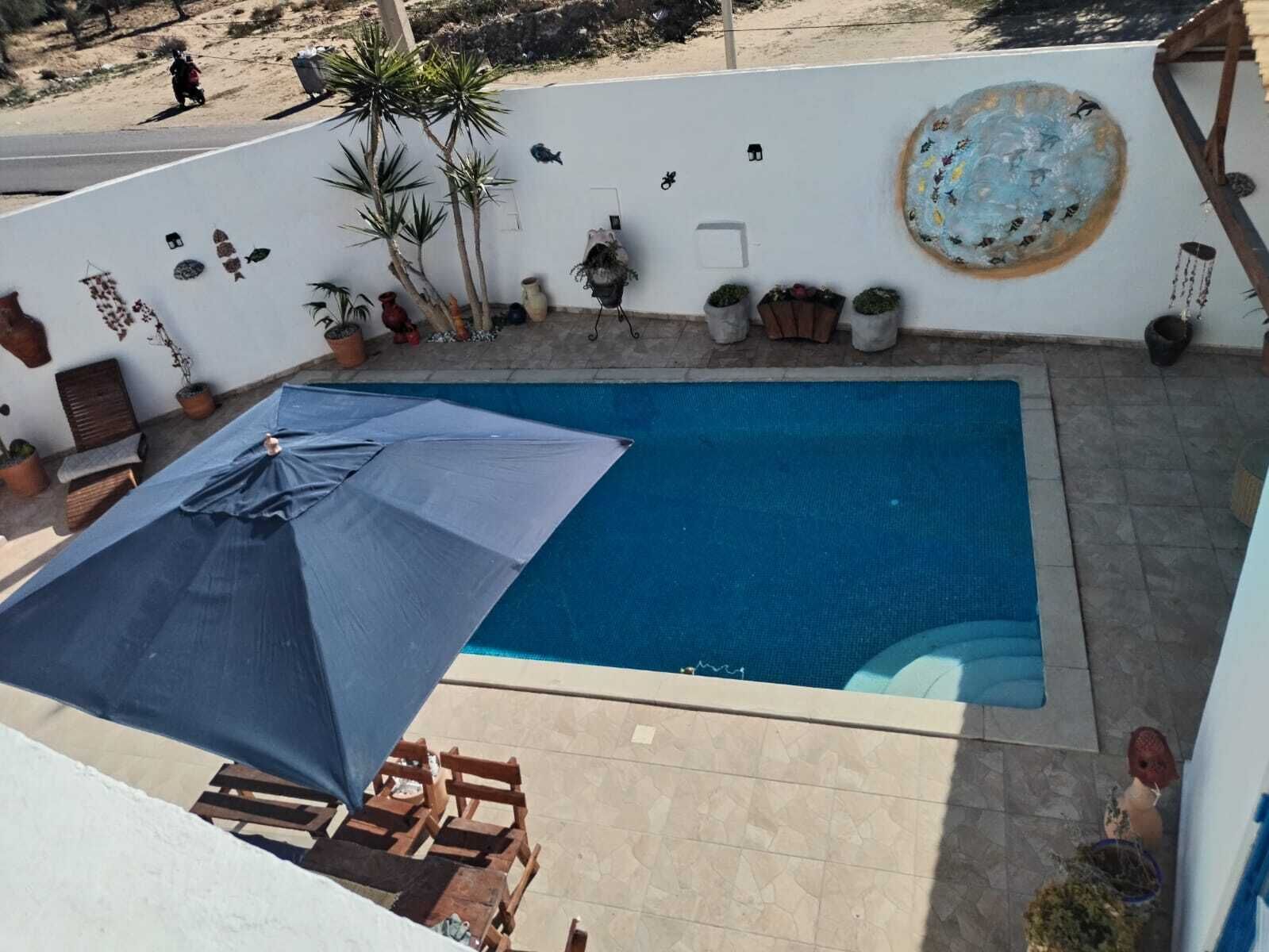 A swimming pool with a blue tarp and wooden chairs, surrounded by potted plants and a white wall with decorative elements.