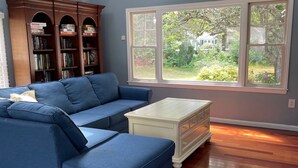 Une pièce comprenant un canapé d’angle, une commode blanche, une bibliothèque en bois et une grande fenêtre donnant sur un paysage verdoyant.