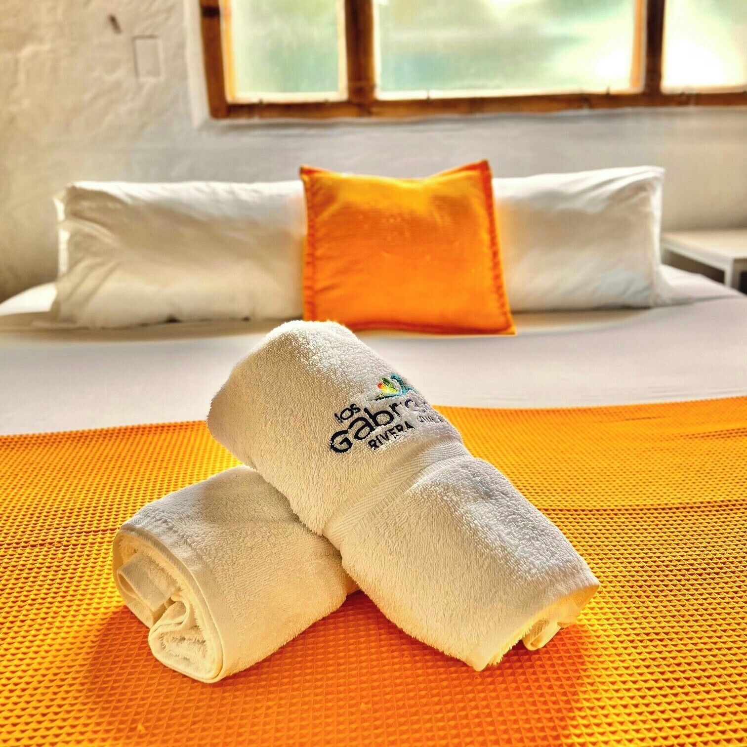 Ein Bett mit weißen und orangefarbenen Kissen, ein aufgerolltes weißes Handtuch und eine gelb strukturierte Bettdecke.