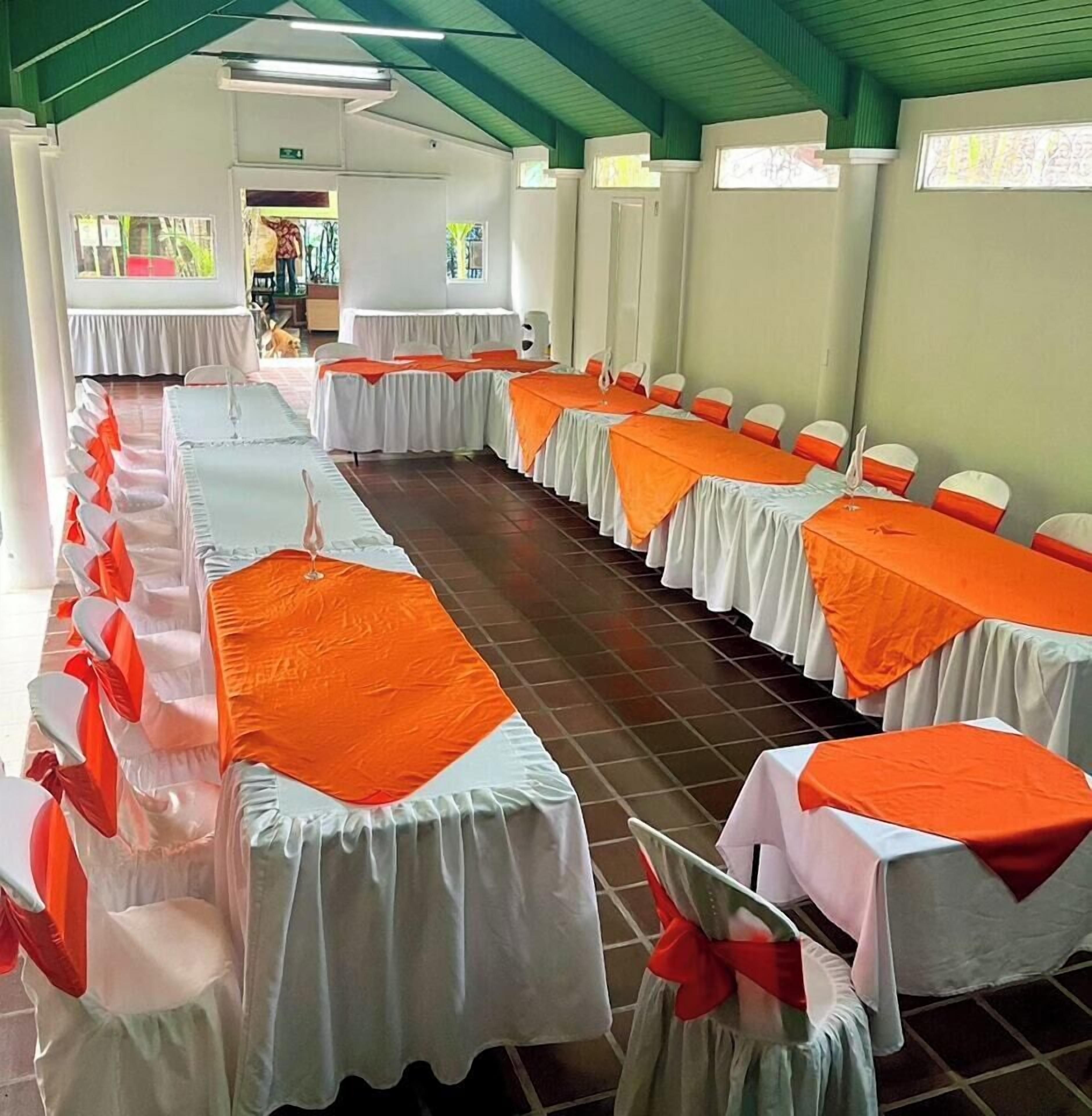 A banquet hall with long tables covered in white tablecloths and orange sashes, arranged in rows.