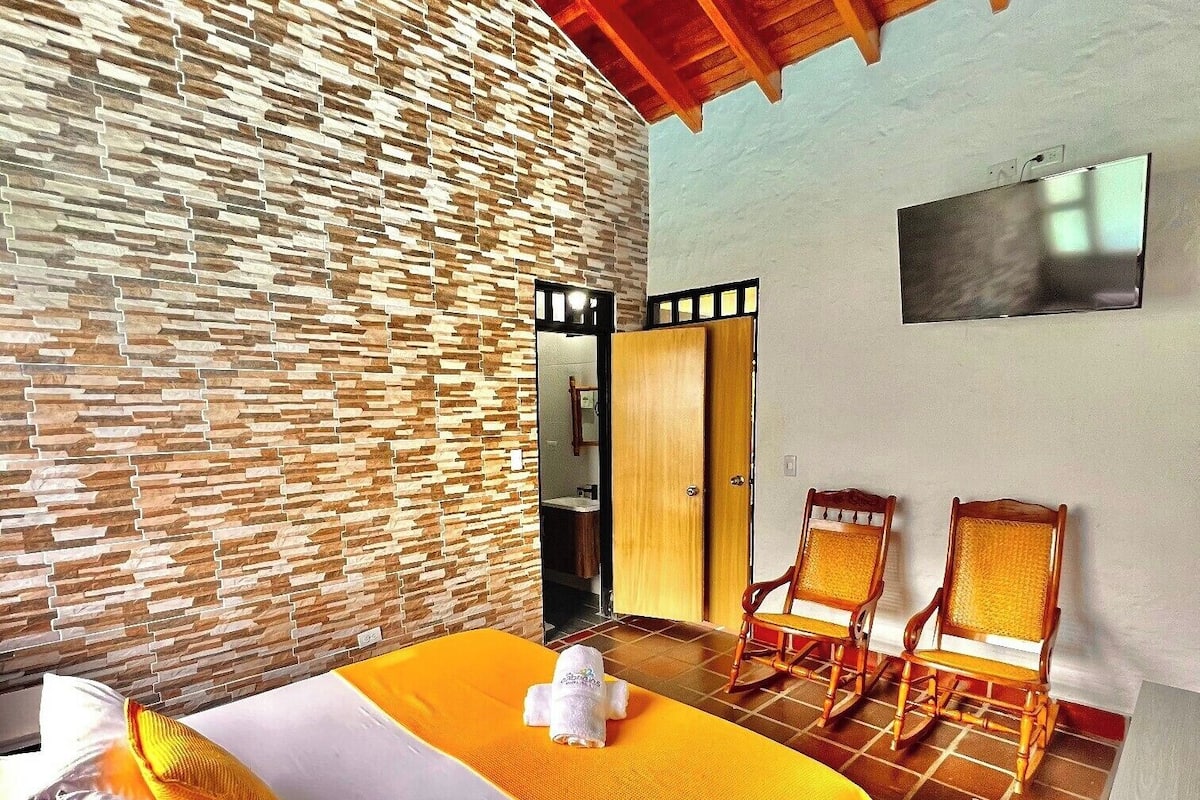 Quarto de hotel com parede de pedra, cama com lençóis brancos, duas poltronas de balanço e teto de madeira.