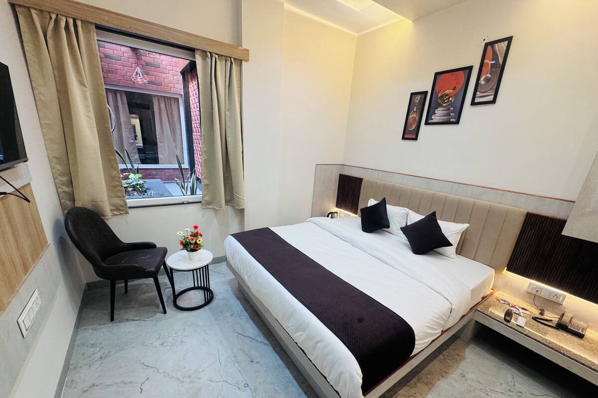 Habitación de hotel con cama, una silla, una pequeña mesa con flores, ventana con cortinas y una obra de arte en la pared.