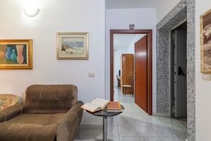 Ein Wohnzimmer mit einem braunen Sofa, einem kleinen Tisch mit Büchern, einem gerahmten Gemälde und einer Tür zu einem anderen Raum.