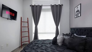 Une chambre à coucher avec un lit, un téléviseur fixé au mur, une échelle et une fenêtre avec des rideaux.