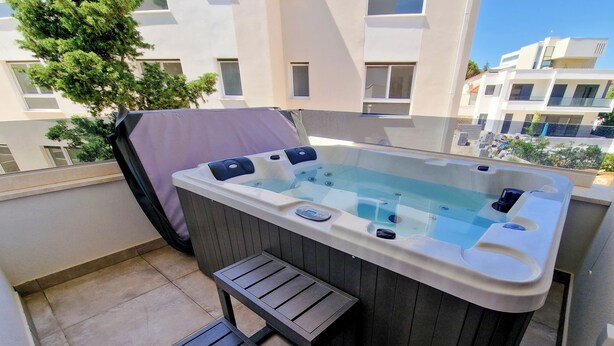Ein Whirlpool mit einer Holzbank und Stufen, einem überdachten Bereich und Blick auf moderne Gebäude.