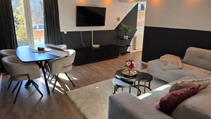 A modern living room with a sofa, dining table, and chairs, a flat-screen TV mounted on the wall, and a staircase leading to an upper level. - Apartment 'Sauerland' with Mountain View, Balcony and Wi-Fi (Willingen (Upland))