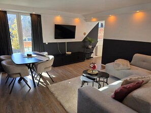 A modern living room with a sofa, dining table, and chairs, a flat-screen TV mounted on the wall, and a staircase leading to an upper level. - Apartment 'Sauerland' with Mountain View, Balcony and Wi-Fi (Willingen (Upland))
