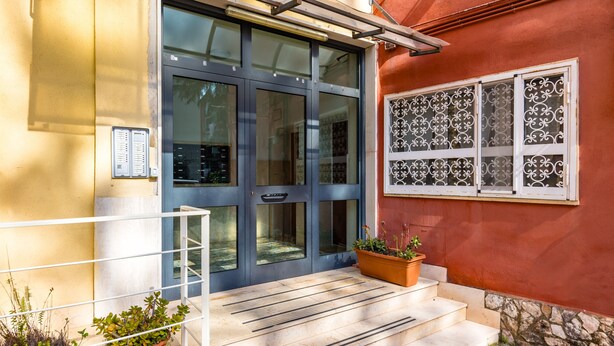 Entrada d'un edifici amb una porta de vidre, una barana metàl·lica i plantes en testos.