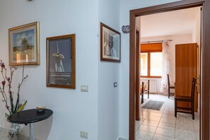 Un pasillo con cuadros enmarcados, una mesita con un cuenco, y una puerta que da a una habitación con una silla y un ventanal.