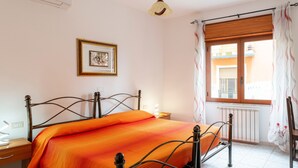 A bedroom with a bed, a bedside table, a lamp, a window with curtains, and a radiator. - Double Room 106 with Air Conditioning (Jerzu)