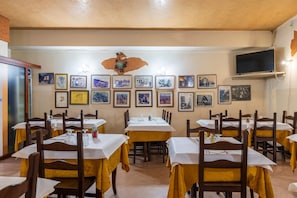 A restaurant with multiple tables, chairs, framed pictures on the wall, and a television mounted on the wall.