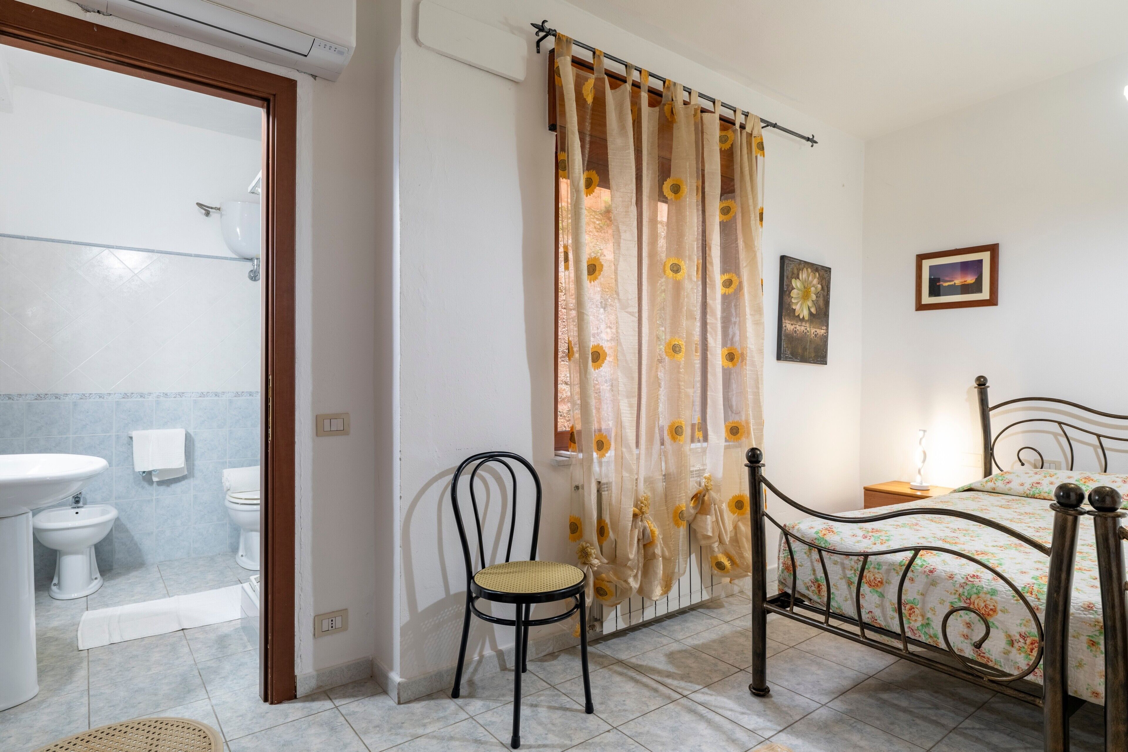 A bedroom with a bed, a chair, a bathroom with a sink and toilet, and a shower area.