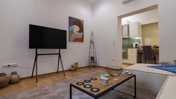 A modern living room with a flat-screen TV, a painting, a ladder, a small table with bowls, and a kitchen visible through an open doorway. - Timeless 1 bedroom Luxury at Panathenaic Stadium (Athina)