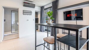 Ein stilvolles Wohnzimmer mit Küchenzeile, einem Esstisch mit Hockern, einem flachbildfernseher an der Wand und einem großen Fenster mit Vorhängen.